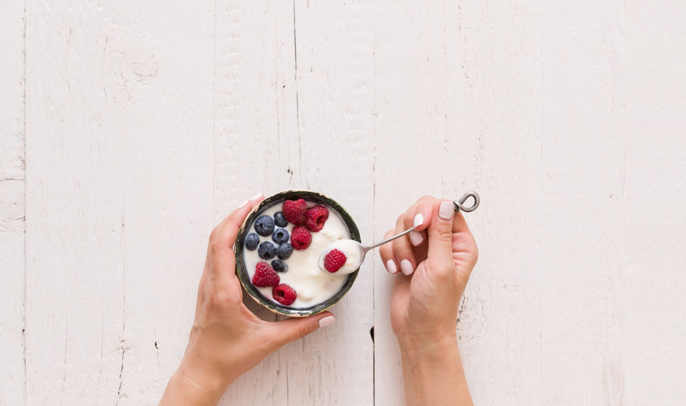 Top view on woman's hands holding small bowl with organic yogurt with blueberries and raspberries on white wooden background